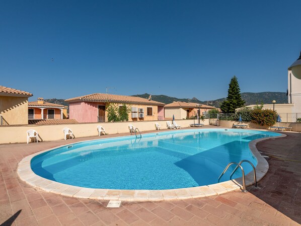 A swimming pool with a clear blue water, surrounded by a tiled deck, and adjacent to buildings with a Mediterranean architectural style.