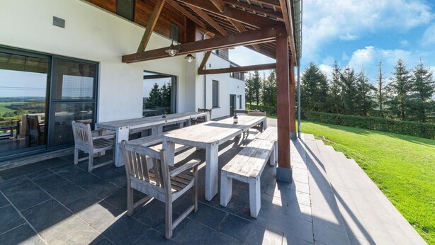 Une terrasse extérieure avec des tables et des bancs en bois, une structure en forme de pergola et une vue sur un paysage verdoyant.
