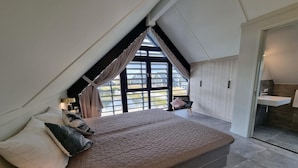 A bedroom with a large bed, a view of the outdoors, a small seating area, and a bathroom.