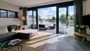 En moderne stue med sofa, sofabord og udsigt til et vandareal samt bygninger gennem store glasdøre.