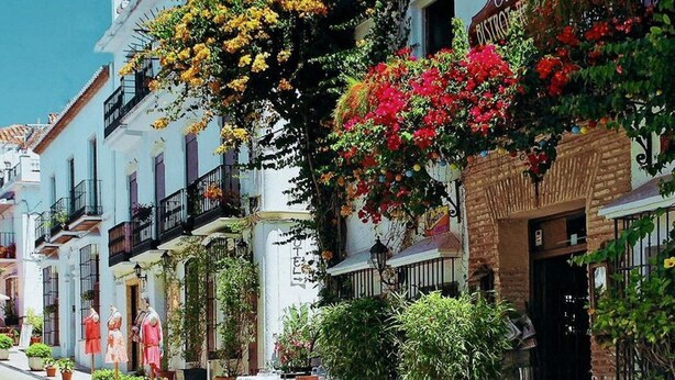 A street with buildings featuring balconies, outdoor seating, and potted plants.