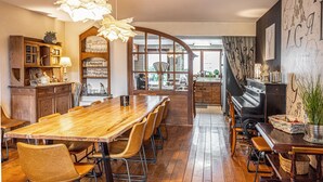 Un comedor amplio con una larga mesa de madera y sillas, un piano y una cocina visible a través de un panel de vidrio.