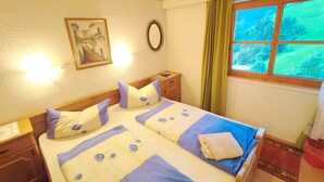 Uma cama de casal com colcha floral, uma mesa de cabeceira, um espelho, um quadro na parede e uma janela com vista para o verde.