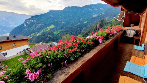 Uma varanda com grade de madeira, uma casa de madeira ao fundo e uma paisagem montanhosa.