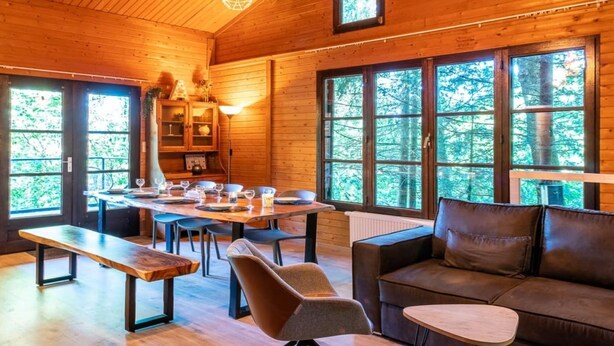 A wooden cabin interior with a dining table set for four, a sofa, and large windows offering a view of the forest. - Chalet in the woods with Nordic bath (Manhay)