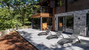 A modern outdoor patio with lounge chairs, a wooden deck, and a stone wall. - Chalet in the woods with Nordic bath (Manhay)