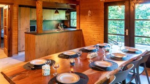 A wooden cabin interior with a dining table set for six, a kitchen area, and a large window with a view of greenery. - Chalet in the woods with Nordic bath (Manhay)