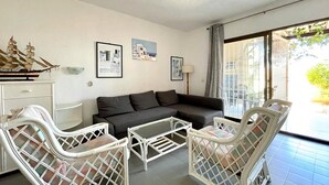 Ein Wohnzimmer mit einer Couch, zwei weißen Korbstühlen, einem Couchtisch und einem Schiffsmodell in einem Schrank. An der Wand hängen drei gerahmte Bilder.