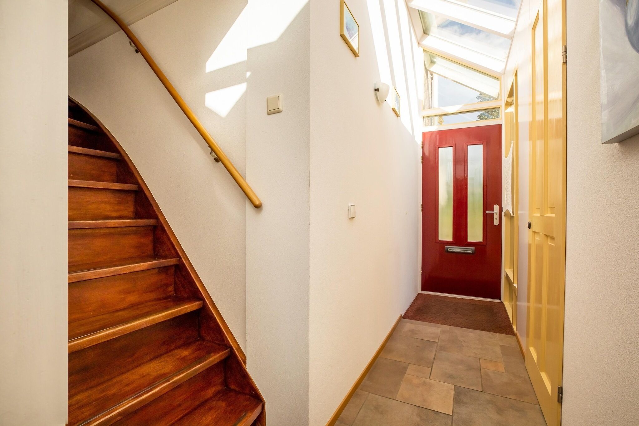 Un couloir étroit, avec un escalier en bois sur la gauche et une porte rouge sur la droite.