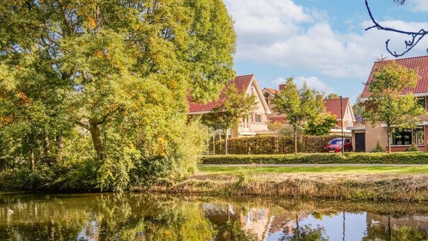 Een rustig meer met weerspiegelingen van bomen en huizen, omgeven door weelderige begroeiing en een heldere lucht.