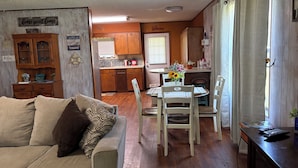 Una sala de estar acogedora con un sofá y una mesa de comedor preparada para cuatro comensales. Se ve una cocina al fondo con armarios de madera y una ventana.