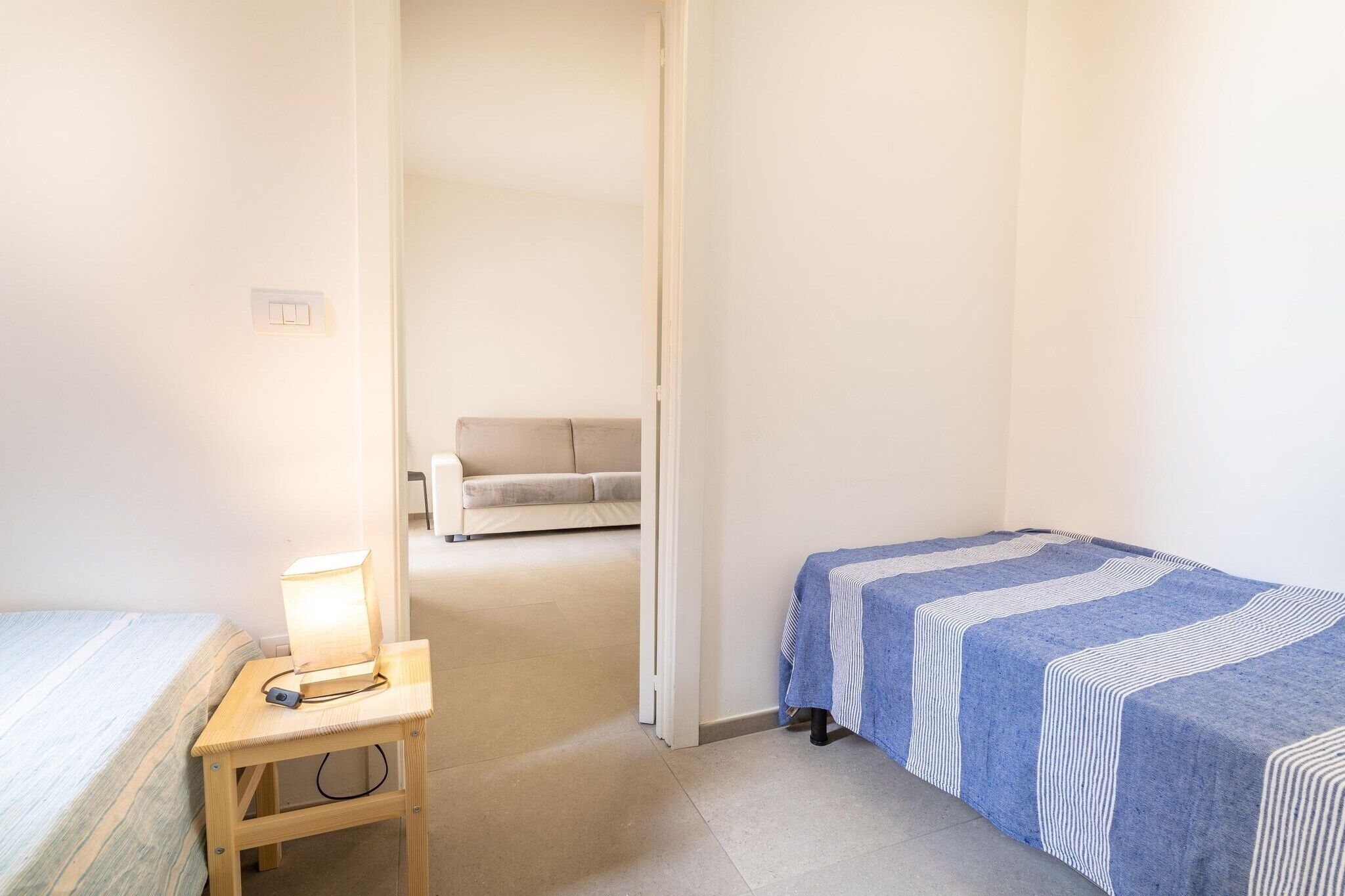 A small, well-lit room with a bed, a bedside table with a lamp, and a sofa in the background.