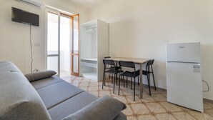 Ein modernes, minimalistisches Zimmer mit einer Couch, einem kleinen Esstisch mit Stühlen, einem Kühlschrank und einem an der Wand befestigten Fernseher.