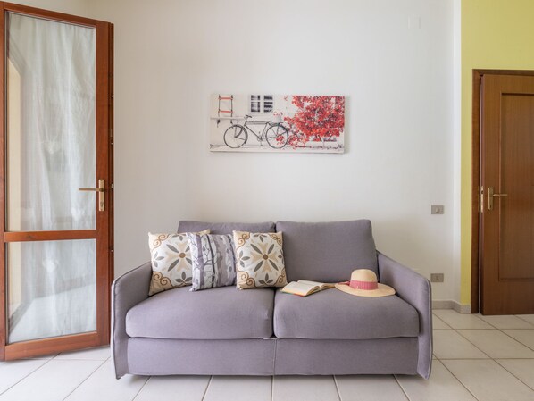 A living room with a grey sofa, patterned cushions, a wall-mounted bicycle artwork, and a door with a wooden frame. - Apartment Iris by Interhome (Villasimius)