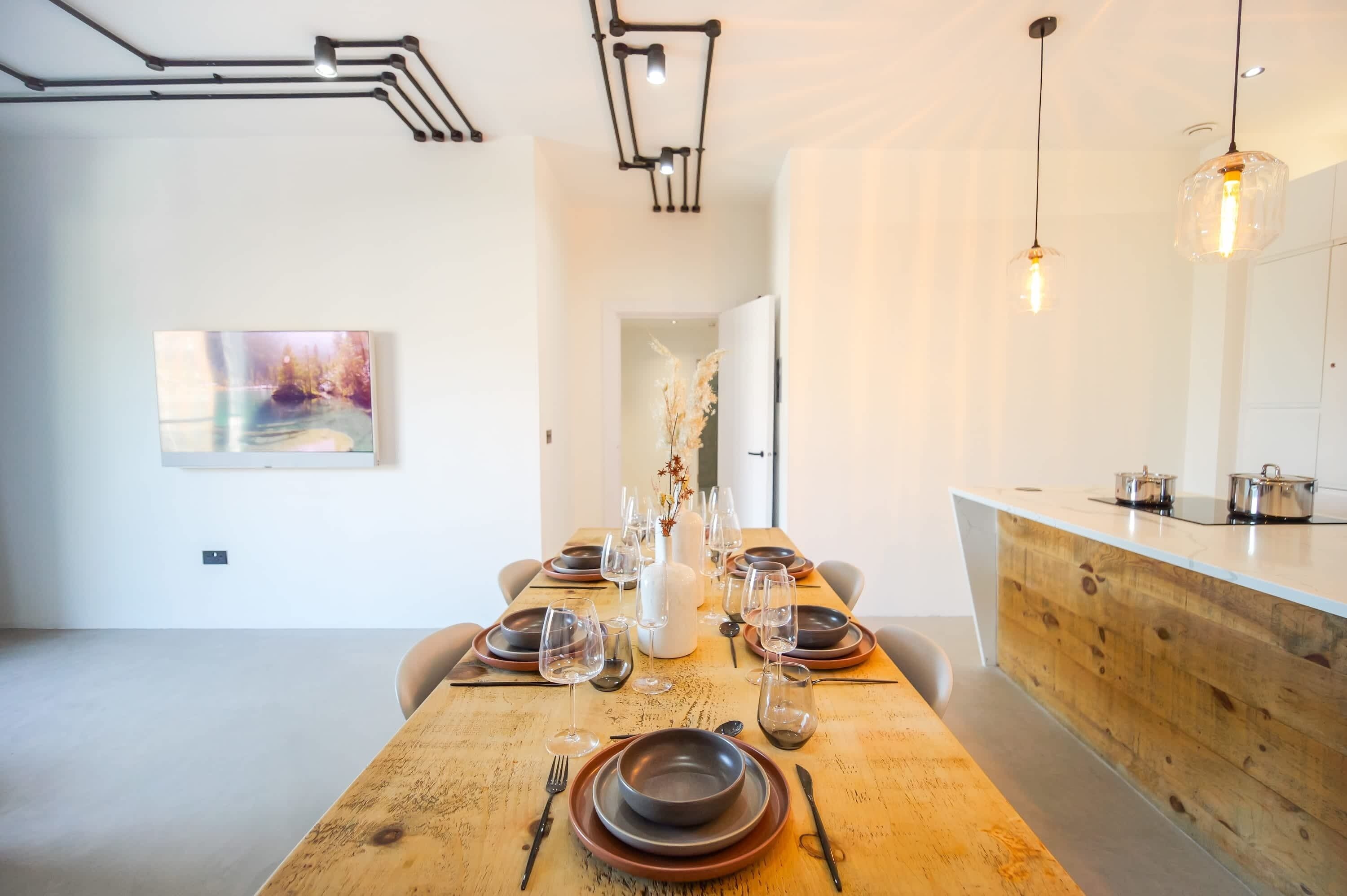 A modern dining area with a wooden table set for a meal, a white vase centerpiece, and pendant lighting.