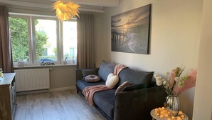 A cozy living room with a dark grey sofa, a small side table with a vase of flowers, and a large window with curtains. - Apartment in Gdańsk near Brzeźno Pier (Gdańsk Brzeźno)