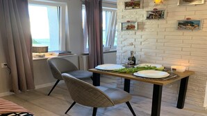A dining area with a table set for two, a chair, a window with curtains, and a brick wall with framed pictures. - Apartment in Gdańsk near Brzeźno Pier (Gdańsk Brzeźno)