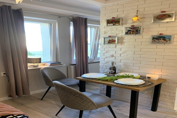 Un coin repas avec une table dressée pour deux personnes, une chaise, une fenêtre avec des rideaux et un mur de briques orné de tableaux encadrés.