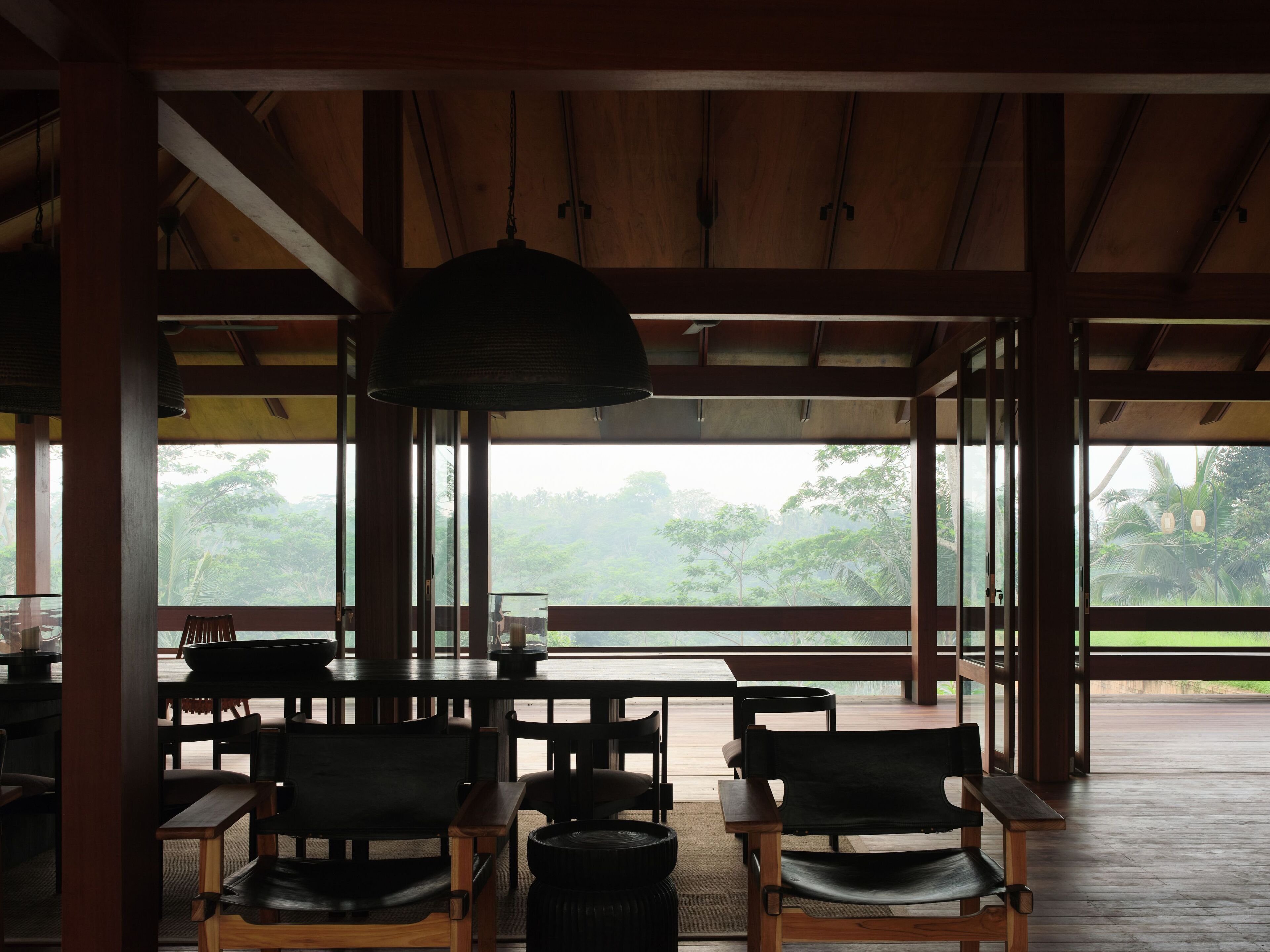 An open-air dining area with wooden tables and chairs, large windows offering a view of greenery.