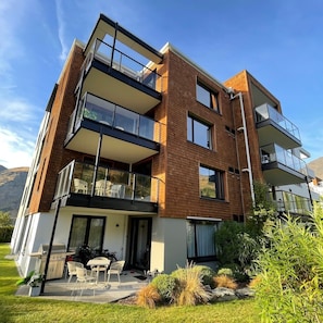 A modern multi-story residential building with balconies, a patio with outdoor furniture, and a mountainous backdrop.