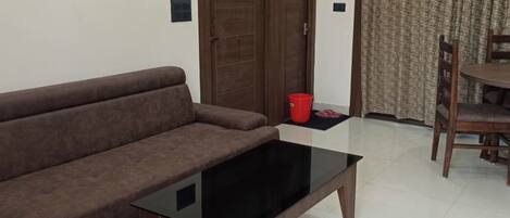 A living room with a brown sofa, a glass coffee table, a wooden door, and a dining area with a table and chairs.