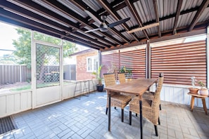 An outdoor patio with a wooden table and wicker chairs, a ceiling fan, and potted plants.
