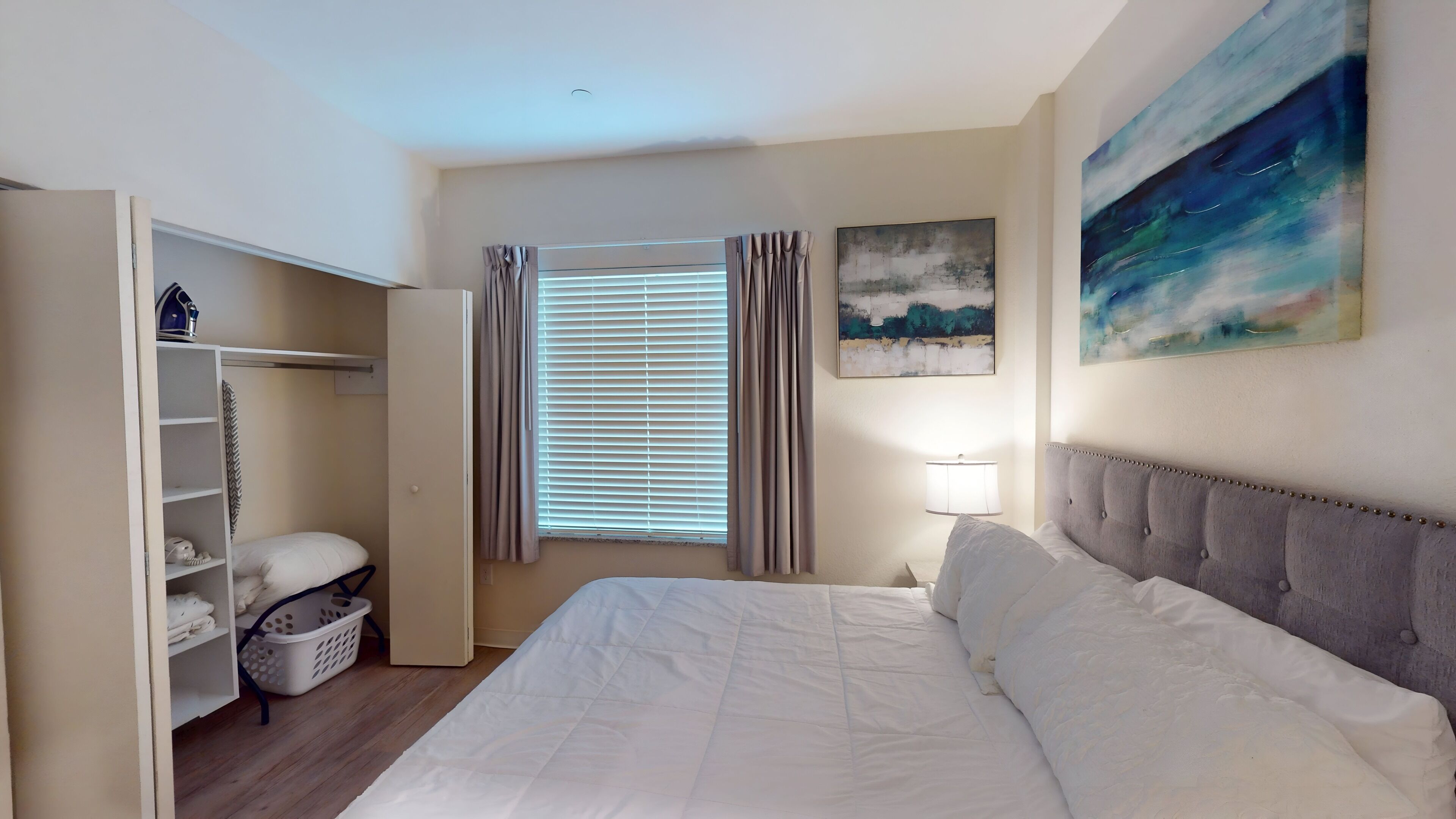 A bedroom with a bed, a wardrobe, a window with curtains, a nightstand with a lamp, and a painting on the wall.