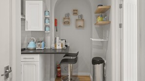 Eine kleine Küche mit weißen Schränken, einem blauen Wasserkocher, einem Toaster und einem Feuerlöscher. Zwei Barhocker und ein Mülleimer sind ebenfalls vorhanden.