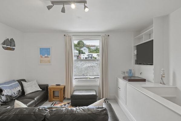 A modern living room with a sofa, a small wooden table, a TV, and a view of the street outside.