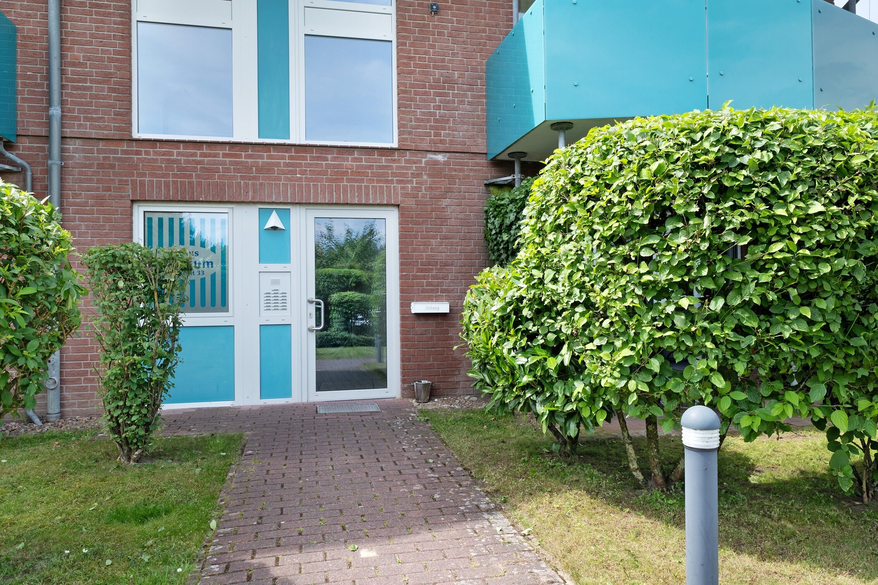 A brick building with a blue door and window, a paved entrance, and a well-maintained hedge.