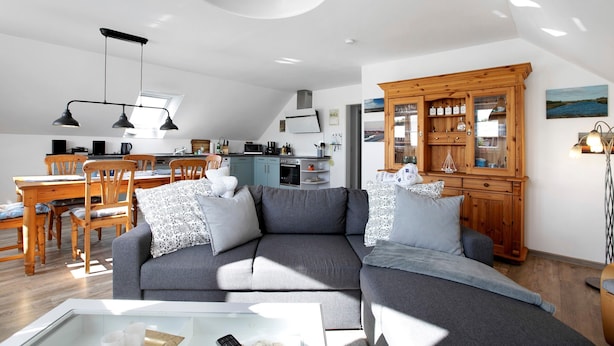 A modern living room with a grey sectional sofa, wooden dining table, and a kitchen in the background. - Apartment 'Ferienwohnung Windrose' with Private Terrace and Wi-Fi (Pellworm)