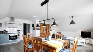A modern kitchen with a built-in oven, a dining area with a wooden table and chairs, a living room with a sofa and TV, and a wooden cabinet in the background. - Apartment 'Ferienwohnung Windrose' with Private Terrace and Wi-Fi (Pellworm)