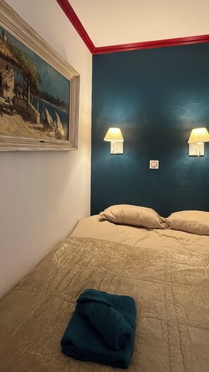 A hotel room with a bed, two wall-mounted lamps, a painting on the wall, and a folded towel on the floor.