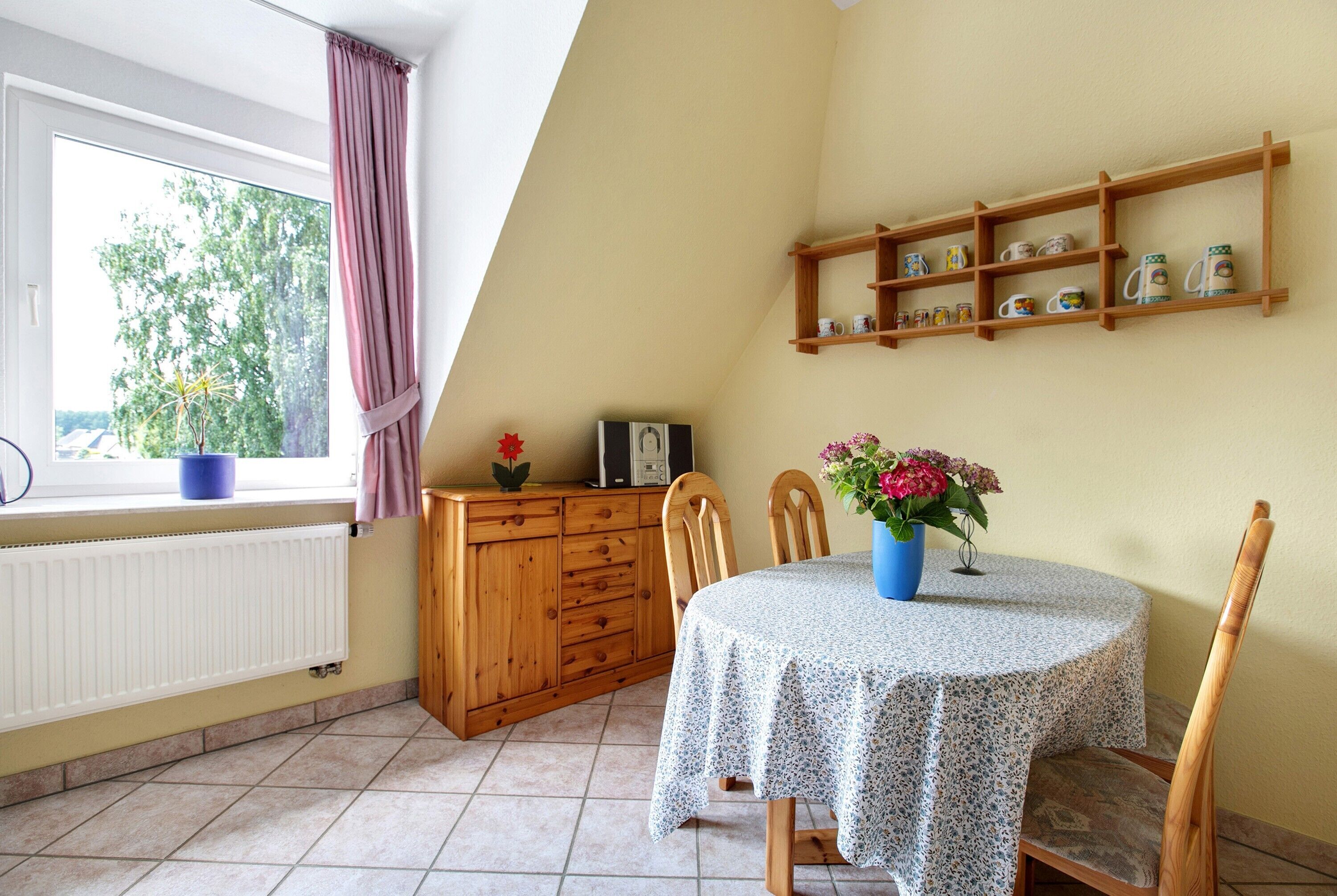 A dining area with a round table, chairs, a window with curtains, a radiator, and a shelf with mugs.