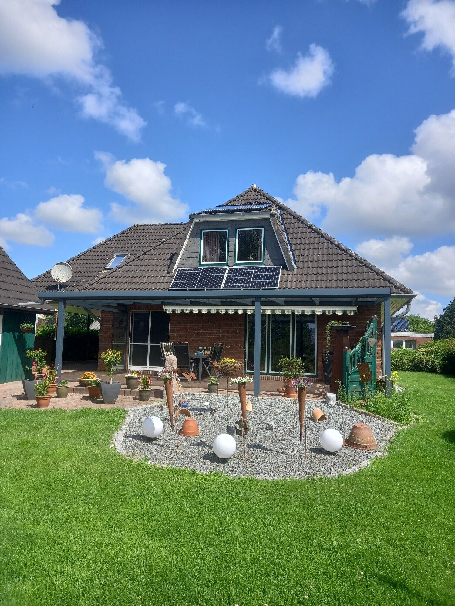 A house with a sloped roof, solar panels, a covered patio, and a garden with potted plants and decorative spheres.