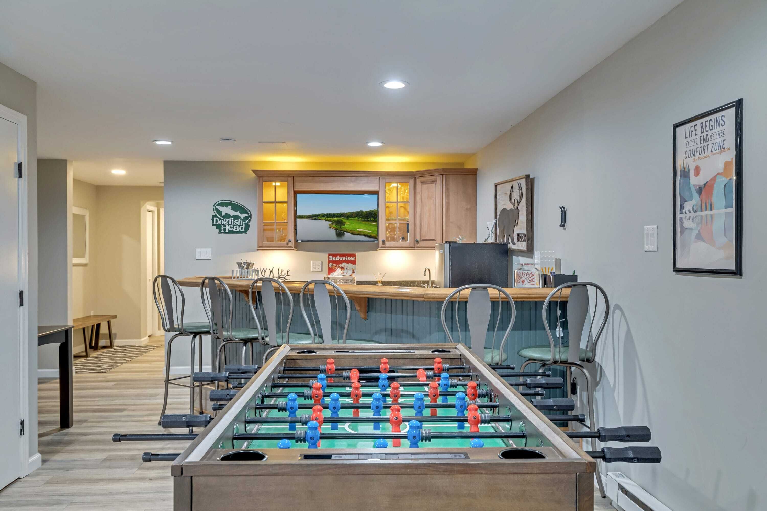 A room with a foosball table, a television, and a kitchen area with a view of a landscape.