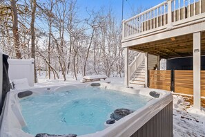 A hot tub surrounded by snow-covered trees, a wooden deck, and a staircase leading to a balcony.