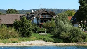 A lakeside house with a balcony, surrounded by greenery and a clear view of the lake.