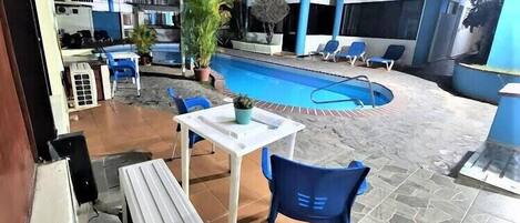 Un espace piscine intérieur avec un sol carrelé, une piscine, des chaises bleues, une table et des plantes en pot.