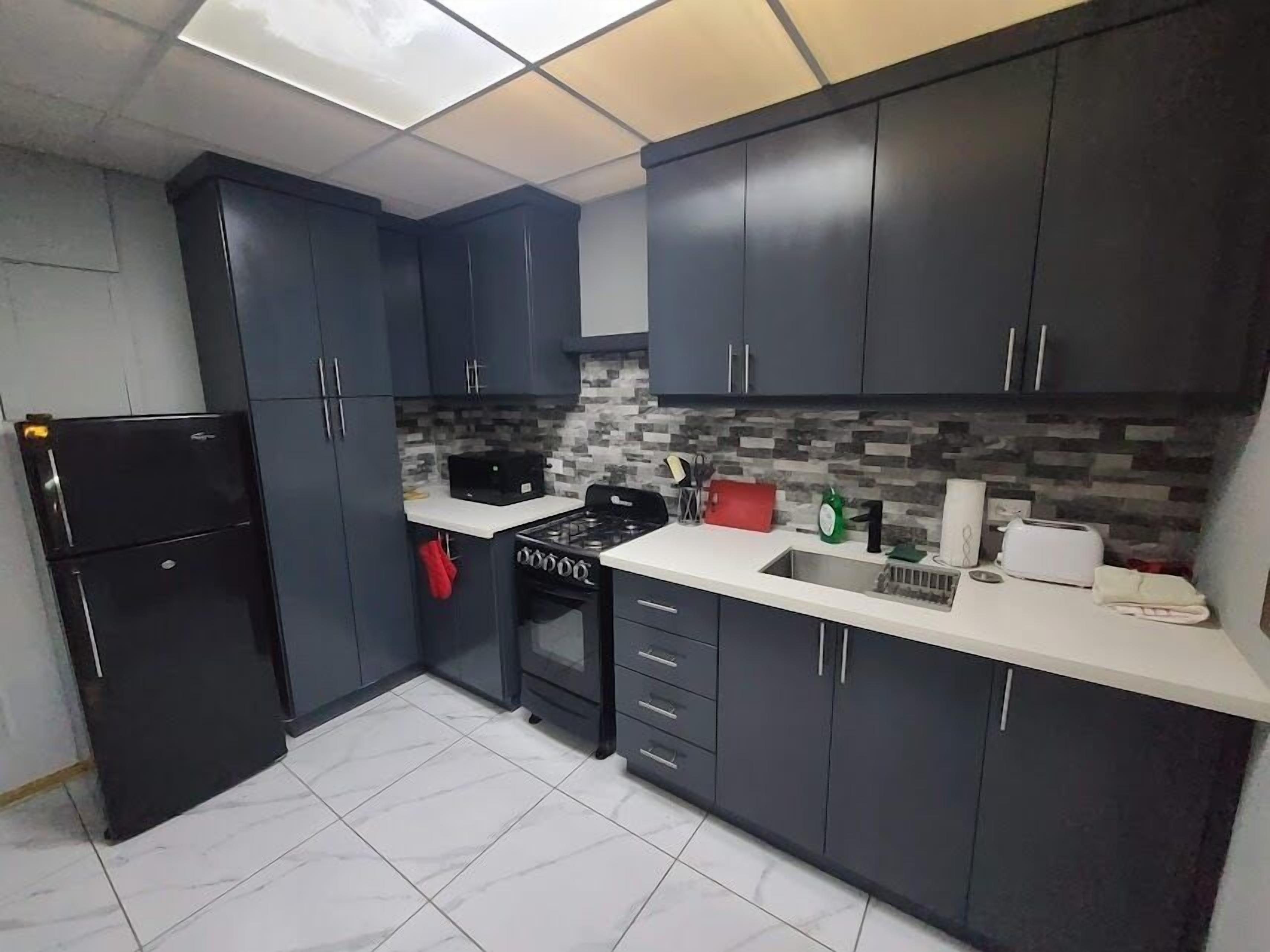 Una cocina pequeña con gabinetes oscuros, un refrigerador negro, una estufa a gas y un salpicadero de azulejos.