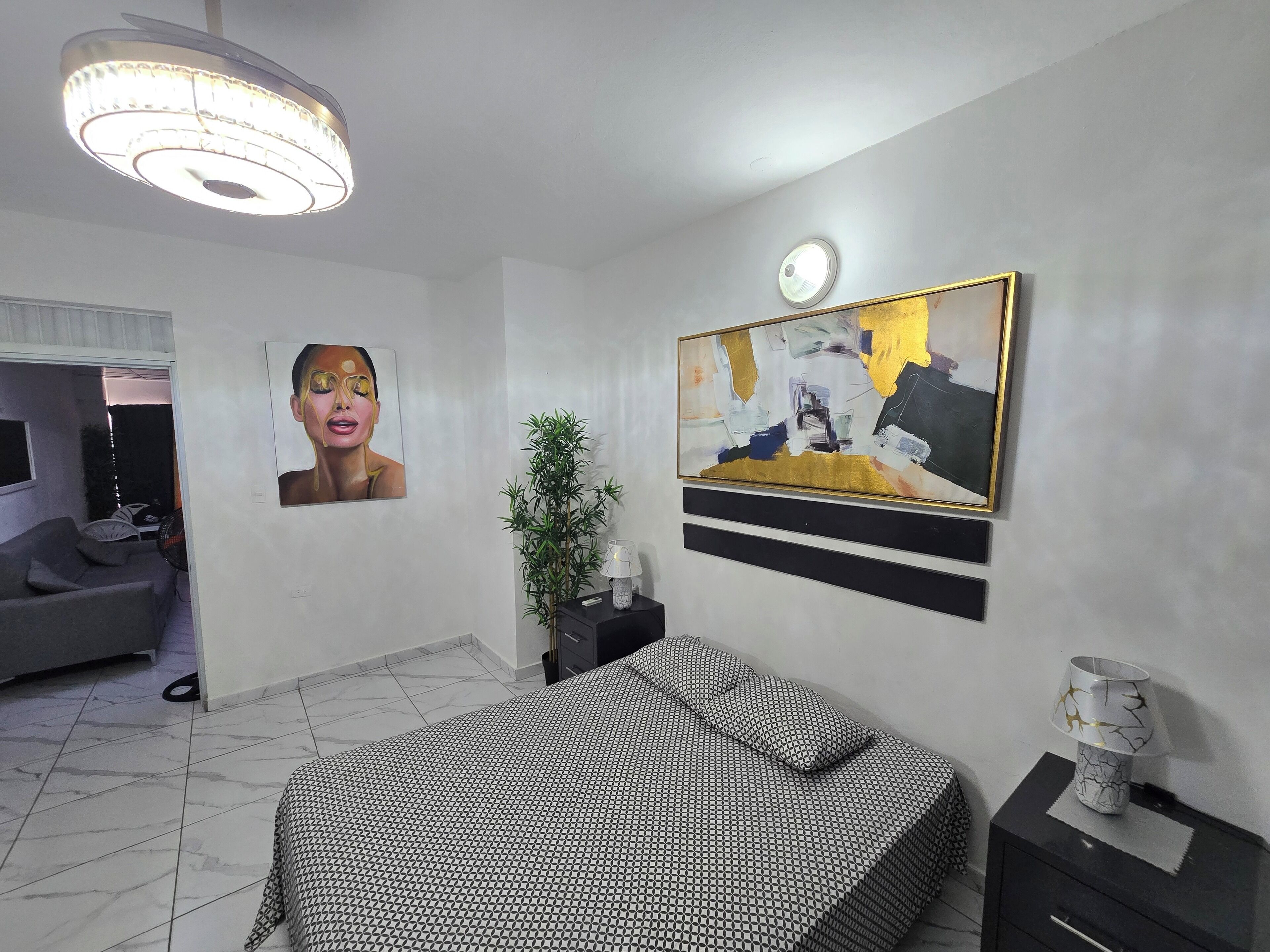 Une chambre à coucher comprenant un lit, une table de chevet, une lampe, un tableau, un luminaire au plafond et une plante en pot.