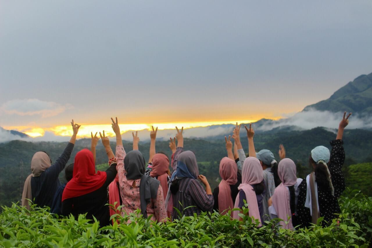 Een groep mensen staat op een heuvel met uitgestoken armen, uitkijkend over een bergachtig landschap tijdens de zonsondergang.
