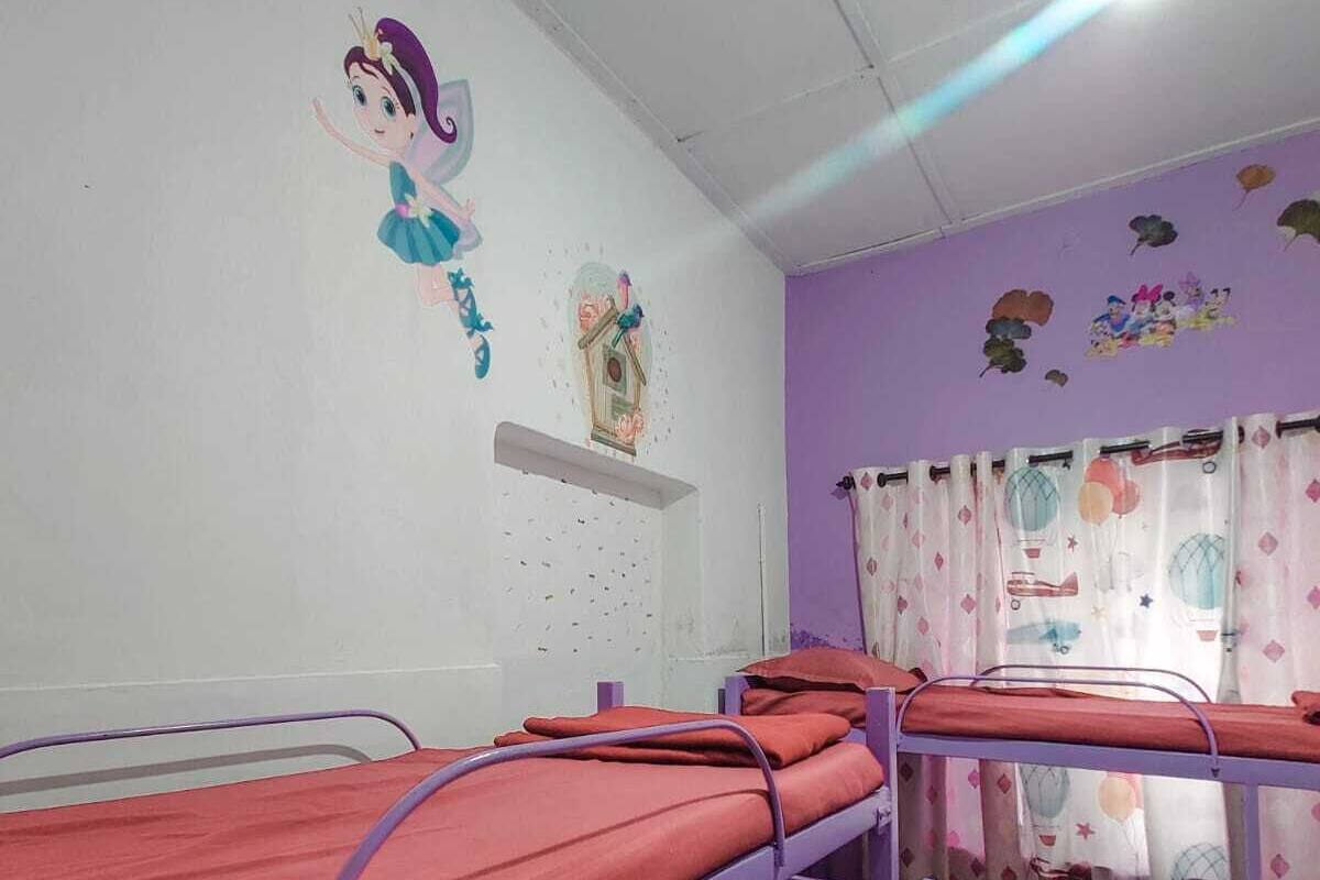 A dormitory room with bunk beds, a small shelf with decorative items, and a window with curtains.