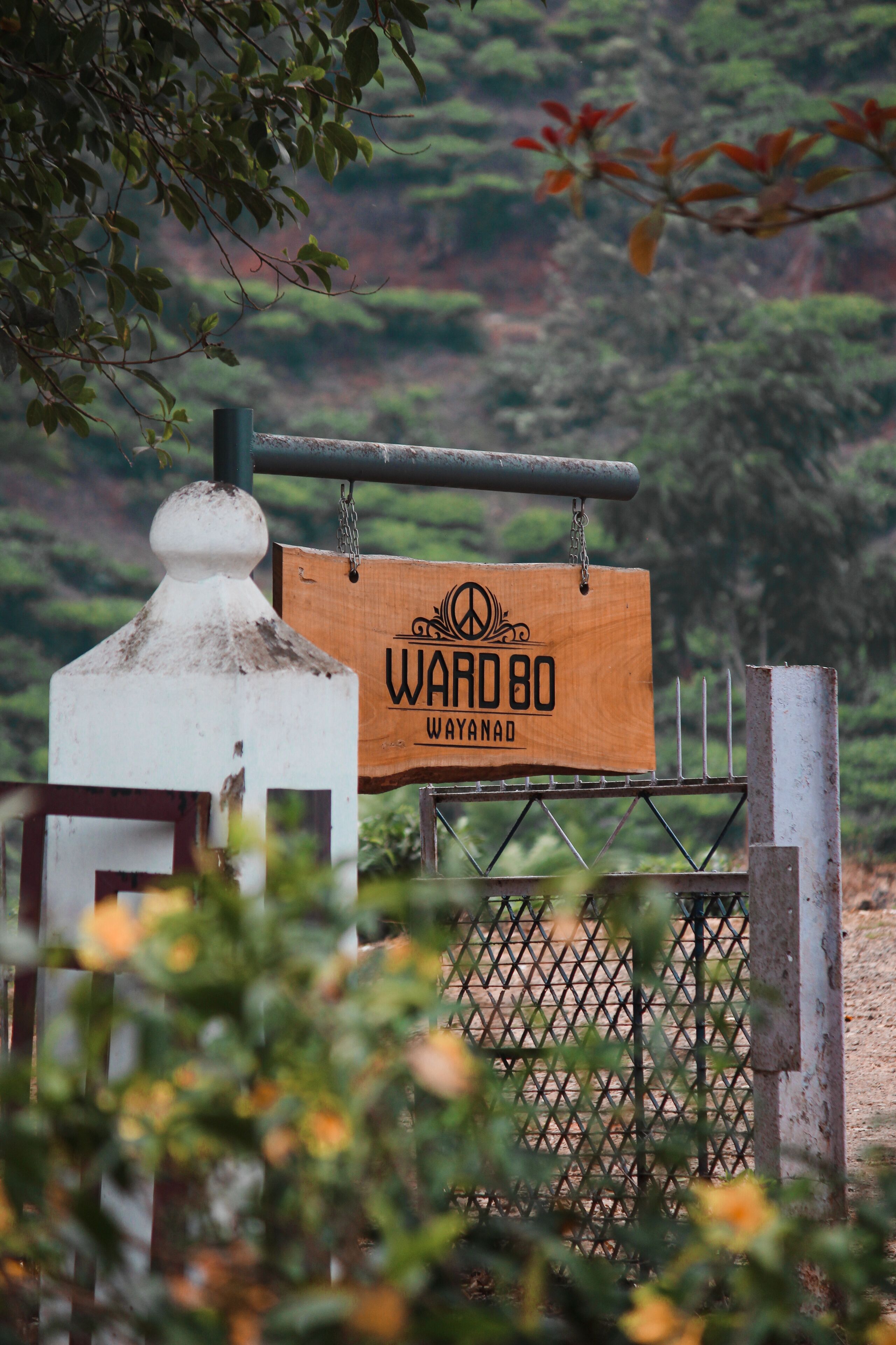 Un letrero de madera con el texto "WARD 80 WAYANAD" colgado de una reja metálica.