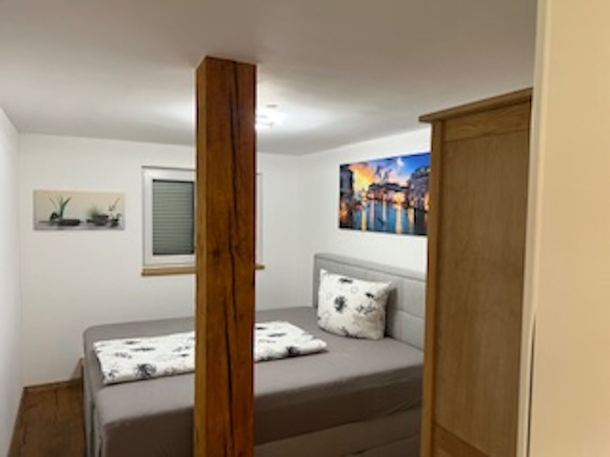 Een slaapkamer met een bed, een raam, een schilderij en een houten kledingkast.