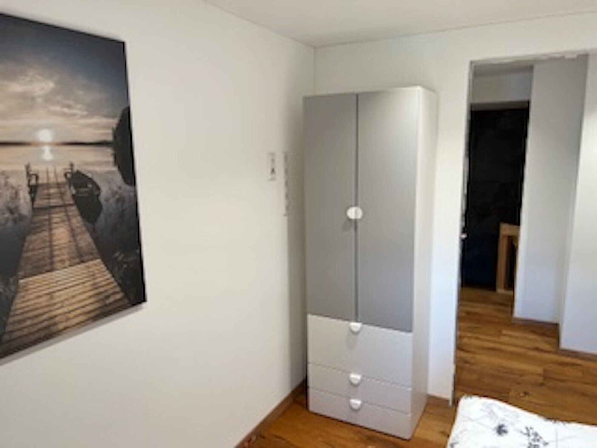 A bedroom with a wooden floor, a white wardrobe, and a painting on the wall.