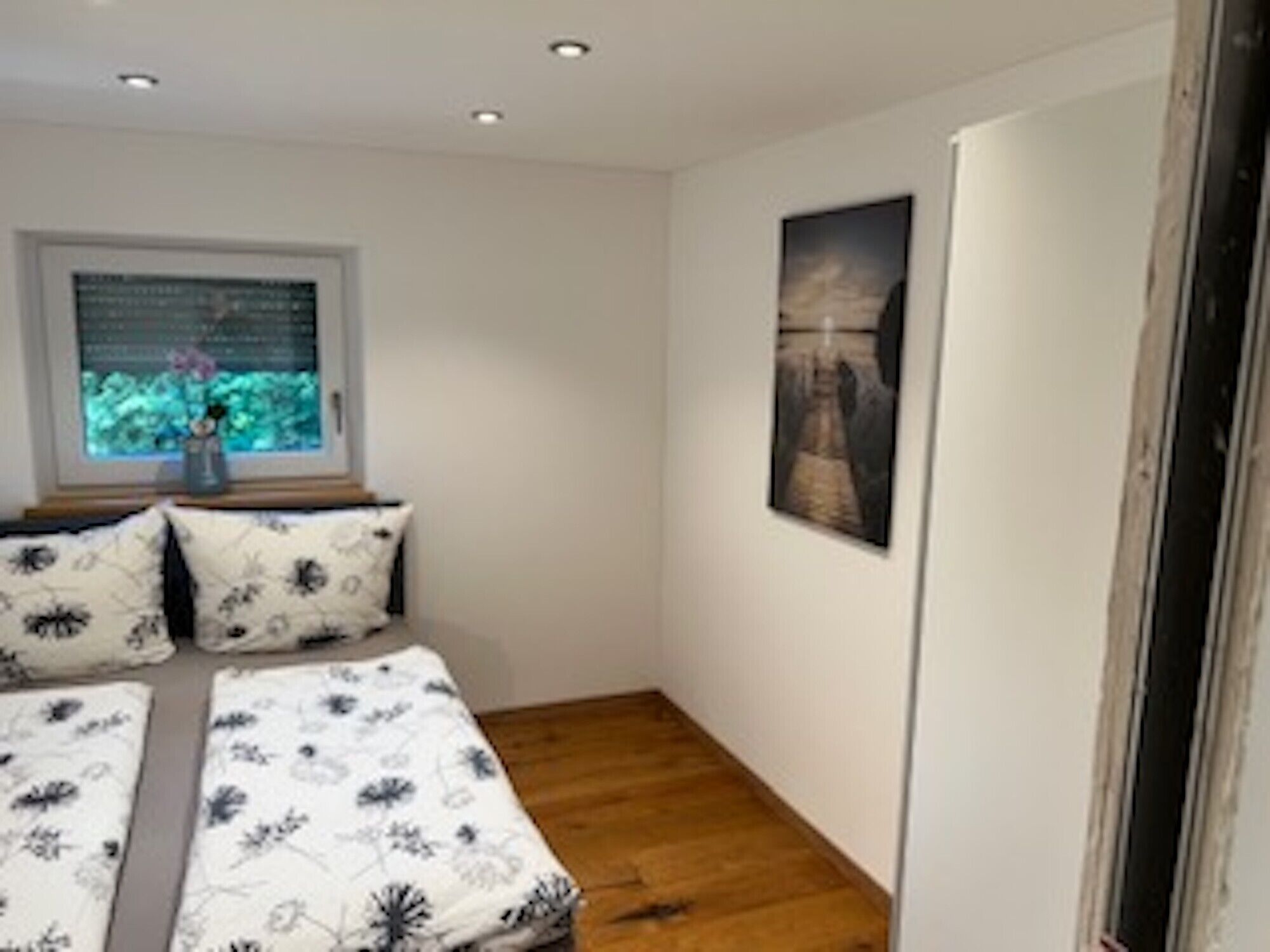 A bedroom with a wooden floor, a window with a view, and a bed with floral bedding.