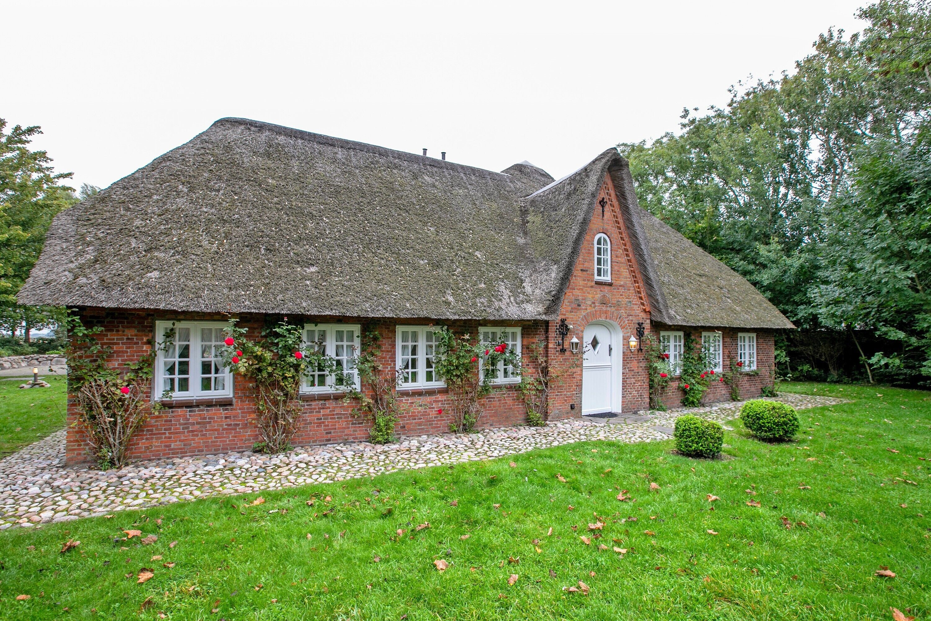 A brick house with a thatched roof, a white door, and a small garden with a hedge and a stone pathway.