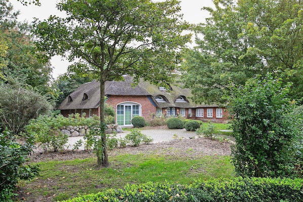 A traditional brick house with a thatched roof, surrounded by lush greenery and a stone wall.
