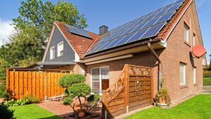 Une maison avec une façade en briques, une clôture en bois, un toit équipé de panneaux solaires et un petit jardin agrémenté d’un banc et de plantes en pot.